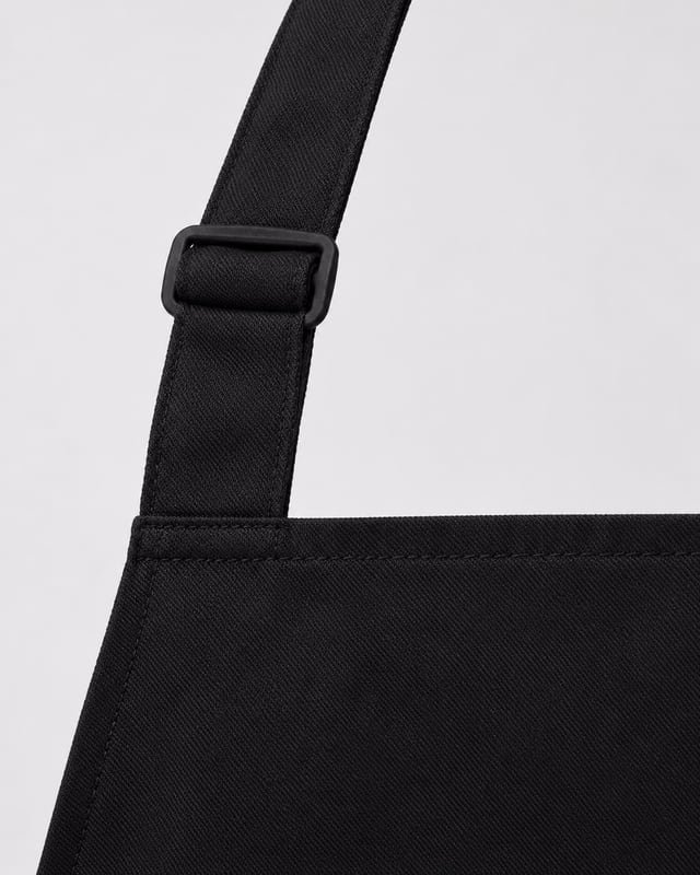 Apron neck-strap adjuster and attachment Reference illustration: Close-up of a black apron neck strap with a rectangular slider and stitched attachment to the bib.