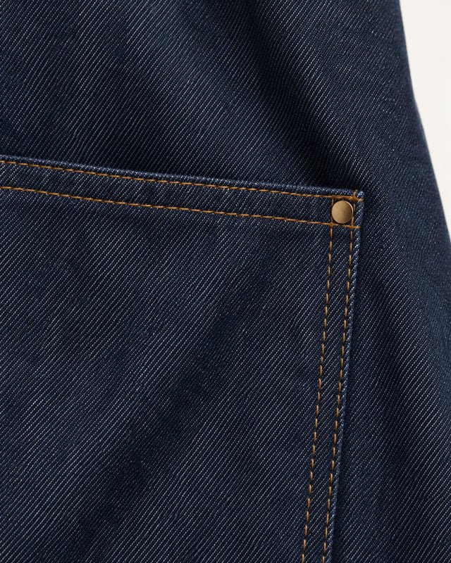 Denim apron fabric and contrast stitching Reference illustration: Close-up of dark blue denim fabric with ochre pocket stitching and a small corner rivet.
