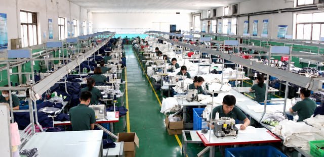 Garment sewing at a manufacturing partner Workers sewing garments along rows of sewing machines at a manufacturing partner, with fabric stacks beside their workstations.